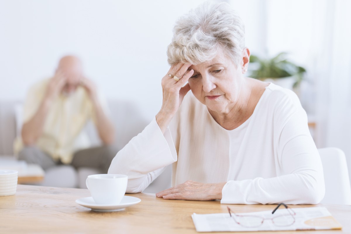 Idős nő tartja a fejét, az agy neurológiai funkcióinak romlása miatt, lehetséges Alzheimer-kór. Idős nő az előtérben, lehajtott fejjel, kezére támaszkodva.